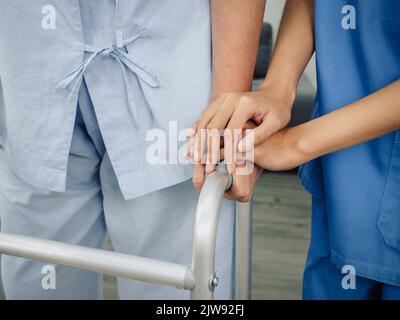 Primo piano delle mani dell'infermiere, assistente in tuta blu che tiene le mani dell'anziano o della donna anziana per il supporto e la cura mentre il paziente anziano cerca di camminare wi Foto Stock