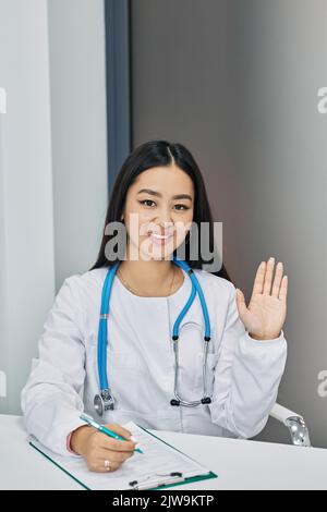 Amichevole medico donna seduto sul suo posto di lavoro indossando uniforme medica e agitando la mano in ospedale Foto Stock