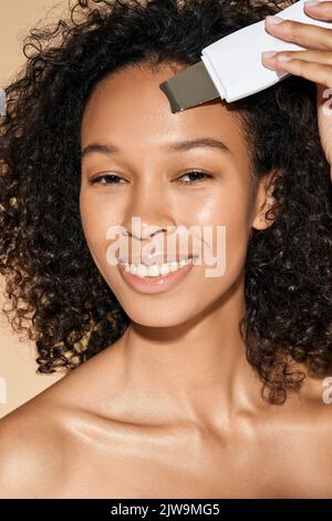 Donna afroamericana sorridente curly che si prende cura della sua pelle con scrubber ad ultrasuoni, rimuovendo l'acne e la pelle di pulizia del suo viso e fronte Foto Stock