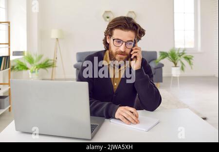 Il solista uomo caucasico Redhead che parla sul telefono cellulare siede alla scrivania dell'ufficio con un computer portatile Foto Stock