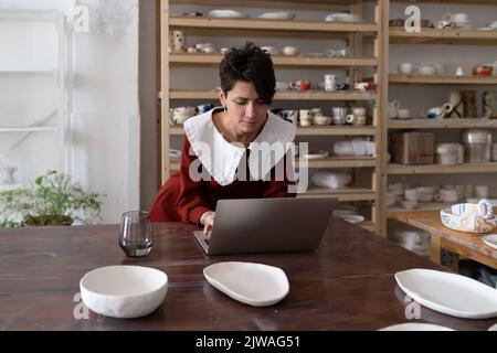 Giovane artigiana femminile che lavora sul computer portatile in ceramica studio, facendo soldi con la ceramica Foto Stock