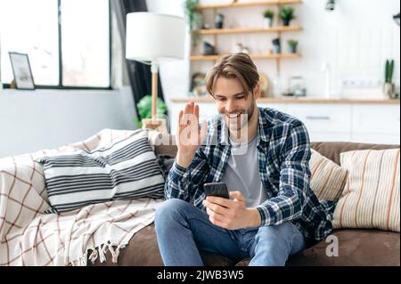 Positivo sicuro amichevole uomo caucasico elegante giovane, indossare abiti casual, sedersi su un divano a casa in accogliente soggiorno, parlare in una videoconferenza da uno smartphone, agitare la mano, sorridere Foto Stock