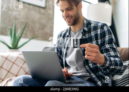 Positivo uomo caucasico defocused acquistare le cose online, utilizzando il laptop e la carta di credito, godendo di shopping in internet, l'acquisto di beni su negozi web, facendo il pagamento a distanza mentre si siede in accogliente soggiorno Foto Stock