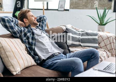 Relax a casa. Soddisfatto positivo calma elegante giovane caucasico, riposante a casa dopo una giornata di lavoro, seduto su un comodo divano, gettando le mani dietro la testa, guardando via, sorridente Foto Stock
