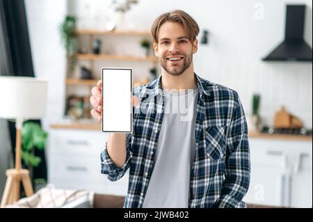 Positivo gioioso caucasico elegante moderno giovane caucasico, in abiti casual, si trova nel soggiorno sullo sfondo della cucina, mostra uno smartphone con uno schermo bianco mock-up, sorriso Foto Stock