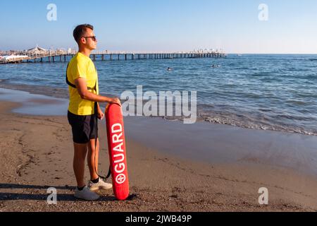 Bagnino maschio sulla spiaggia mediterranea che guarda le persone in acqua. Sicurezza durante il nuoto, bella brunette maschio bagnino sulla spiaggia. Foto Stock