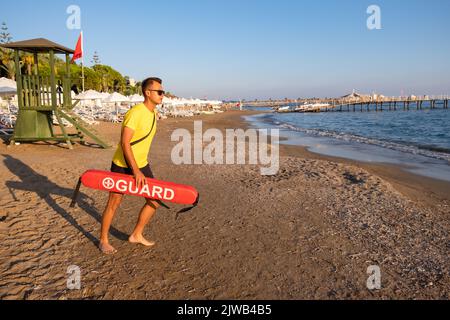 Bagnino maschio sulla spiaggia mediterranea che guarda le persone in acqua. Sicurezza durante il nuoto, bella brunette maschio bagnino sulla spiaggia. Foto Stock