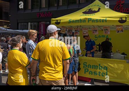 Le persone che indossano camicie e bandiere brasiliane godono di una fiera di strada sulla Sesta Avenue in sostituzione della festa brasiliana a New York. Per il terzo anno consecutivo, il BR Day New York, il più grande partito brasiliano al di fuori del Brasile, non si svolge. João de Matos, presidente del BR Day NY, afferma che l'evento a cui hanno partecipato 1,5 milioni di persone è stato sospeso a causa di una combinazione di fattori, tra cui il recente scoppio di monkeypox a New York; recenti manifestazioni a causa delle elezioni presidenziali in Brasile il prossimo ottobre; e questioni logistiche e di trasporto aereo. Foto Stock