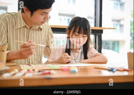 Una ragazza asiatica piccola cute sta concentrando l'immagine della pittura con un acquerello, imparando l'arte con il suo insegnante privato. Concetto di svago per bambini Foto Stock