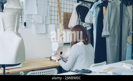 Vista posteriore di una giovane donna designer di abbigliamento che lavora con macchina da cucire seduto alla scrivania dello studio. Sono visibili articoli da cucire, manichino da sartoria e abiti da donna semifiniti. Foto Stock