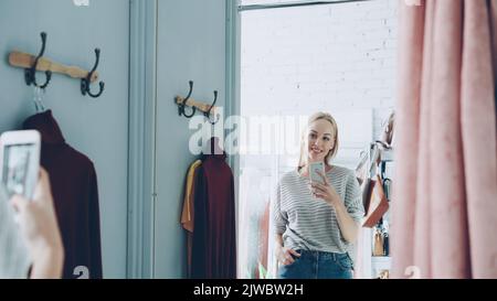 Bella donna bionda sta facendo selfie specchio con telefono intelligente mentre si trova in piedi in una bella stanza vestito in boutique di lusso vestiti. Sta posando, muovendosi e sorridendo con noncuranza. Foto Stock