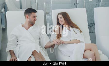 Bell'uomo barbuto e la sua bella ragazza che indossa accappatoi sono seduti in sedie nella sala relax in day spa, bere cocktail e parlare. Concetto di rapporto e benessere. Foto Stock