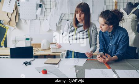 Le giovani donne attraenti che lavorano nell'industria di disegno stanno confrontando gli schizzi e parlando di loro mentre lavorano nel negozio moderno di sarto. Concetto di lavoro di squadra produttivo. Foto Stock