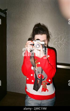 Foto analogica verticale per interni di una donna caucasica dai capelli scuri irriconoscibile di mezza età in un maglione rosso natalizio invernale con una renna sorridente scattando una foto con una fotocamera analogica. Foto di alta qualità Foto Stock