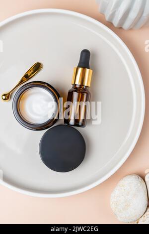 Set di cosmetici naturali anti-età per la cura della pelle su vassoio in ceramica su fondo a baige. Olio viso di bellezza, olio essenziale e crema viso in vetro scuro senza marchio Foto Stock