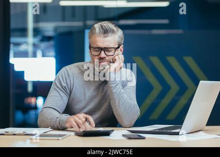 Sconvolto e deluso senior e maturo finanziatore, uomo che lavora all'interno della costruzione di uffici, il calcolo del rapporto finanziario con calcolatrice, grigio haired imprenditore investitore in occhiali pensando pratiche Foto Stock