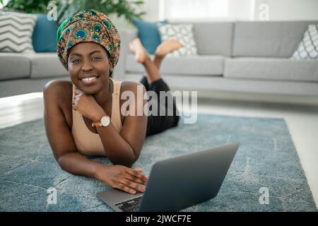 Sorridente giovane ragazza africana am seduta con le gambe incrociate sul tappeto a casa utilizzando il computer portatile e guardando in macchina fotografica Foto Stock