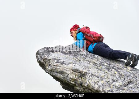 Donna escursionista con zaino rosso si siede al bordo di una scogliera e guarda verso il basso stupito e spaventato, isolato dalla nebbia. Escursioni in Slovacchia alta Tatra montagne. Foto Stock