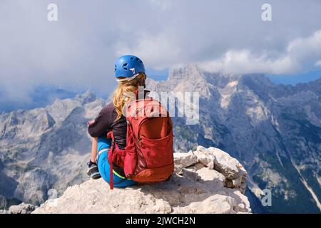 Una donna escursionista con zaino si siede sulla cima del Triglav e guarda le montagne circostanti nelle Alpi Giulie, Slovenia. Estate, attivo, attivo Foto Stock