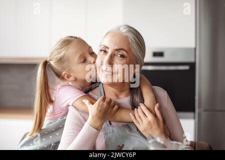 Ritratto di sorrida nipote europea bacia guancia di anziana nonna in grembiule Foto Stock