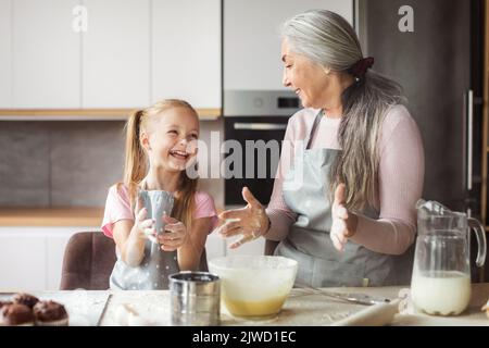 La piccola nipote europea soddisfatta e la nonna matura in grembiuli fanno l'impasto con la farina, divertitevi insieme Foto Stock