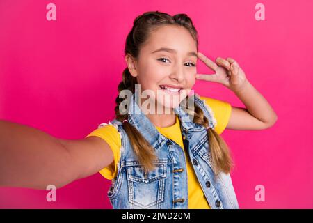 Foto di cool piccola ragazza bionda fare selfie mostra v-segno indossare giallo t-shirt jeans giubbotto isolato su sfondo fucsia Foto Stock