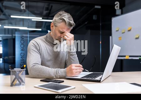 Uomo d'affari anziano dai capelli grigi overstanco che lavora sul laptop, uomo maturo che indossa gli occhiali ha dolore negli occhi, capo che lavora in azienda all'interno di un moderno edificio per uffici. Foto Stock