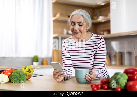 Ritratto di donna anziano sorridente utilizzando smartphone e bere caffè in cucina Foto Stock