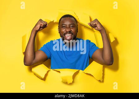 Un uomo afro-americano positivo che si acclama con pugni aggrappati, festeggia la vittoria, in piedi nel buco lacerato di carta gialla Foto Stock