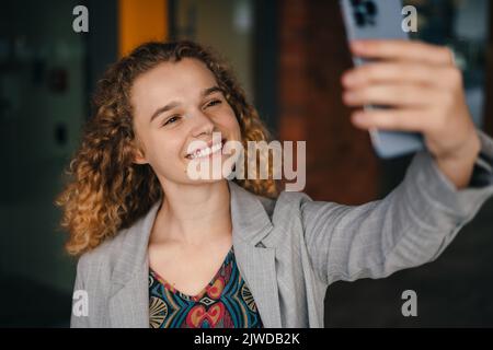 Sorridente overjoyed divertimento giovane studente donna ricci-capelli con giacca facendo selfie sparato sul cellulare all'aperto. Moda femminile. Ritratto di bellezza Foto Stock