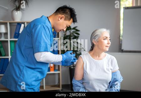 Grave adulto asiatico uomo medico fare l'iniezione a vecchia donna paziente in clinica ufficio interno Foto Stock