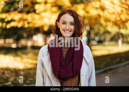 Felice donna europea millenaria rossa in impermeabile con sciarpa Godetevi il bel tempo, tempo libero nel parco cittadino Foto Stock