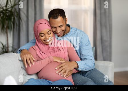 Romantico nero incinta sposi musulmani che si rilassano insieme sul divano in soggiorno Foto Stock