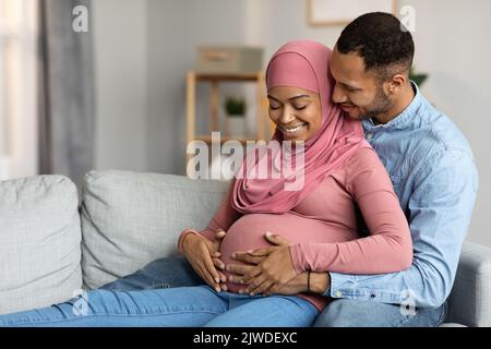 Marito amorevole abbracciando moglie musulmana incinta mentre si rilassa sul lettino a casa Foto Stock