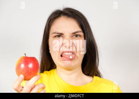 Una donna con un grimace delirato di pillole sulla sua lingua tiene una mela. Rifiuto di integratori alimentari sintetici e vitamine. Foto Stock