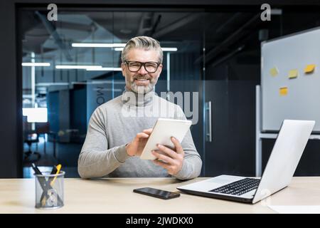 Ritratto del leader di successo del team di programmatori, ingegnere maturo dai capelli grigi che prova la nuova app sul computer tablet, uomo d'affari che lavora all'interno di un moderno edificio per uffici, sorridendo e guardando la fotocamera Foto Stock