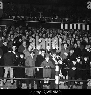 1960s, storico, tifosi di calcio in piedi insieme sulle terrazze per una partita serale ... cappotti, cappellini piatti per gli uomini, duffel cappotti e cappelli bobly per i giovani, non una replica camicia in vista! Foto Stock