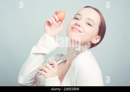 La ragazza si trova su sfondo bianco, frusta a palloncino e agg e cuochi, corsi di cucina, cuochi scolastici. La ragazza grimaces, mostra emozioni, è sorpreso, Foto Stock