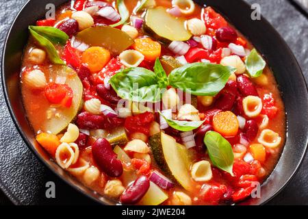 minestrone con fagioli rossi, zucchine, garbanzo, conchiglie di pasta, formaggio, pomodoro, carote e basilico in recipiente nero, primo piano Foto Stock