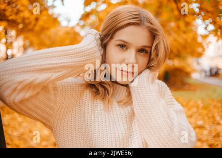 Ritratto fotografico di una ragazza in un maglione bianco, la bionda tiene il collo del maglione e guarda la fotocamera. Bella donna nel parco in autunno. AUT Foto Stock