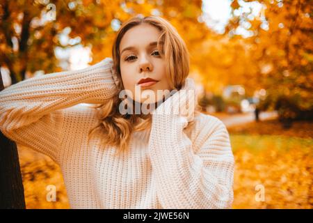 Ritratto fotografico di una ragazza in un maglione bianco, la bionda tiene il collo del maglione e guarda la fotocamera. Bella donna nel parco in autunno. AUT Foto Stock