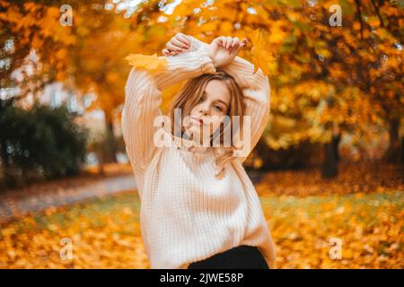 Ritratto fotografico di una ragazza in un maglione bianco, la bionda tiene le foglie in mano. Bella donna nel parco in autunno. Moda e stile autunnali. Caldo Foto Stock