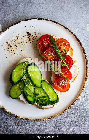 Vista dall'alto di due fette di pane tostato con formaggio cremoso, cetriolo, pomodoro, rucola e semi di sesamo Foto Stock