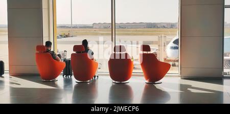 Un paio di giovani passeggeri si sono seduti in comode sedie moderne che si affacciavano dalla finestra di una sala partenze dell'aeroporto Foto Stock