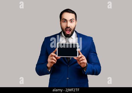 L'uomo bearded che mostra lo schermo vuoto del tablet per adv, ha un volto incredibile, guardando la fotocamera con la bocca aperta, indossando tuta di stile ufficiale. Studio in interni isolato su sfondo grigio. Foto Stock