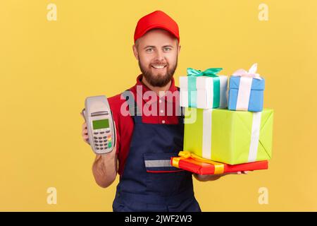 Ritratto di un corriere felice positivo che indossa tute blu che tengono una pila di scatole di presentazione e pos terminal per il pagamento online. Studio al coperto isolato su sfondo giallo. Foto Stock