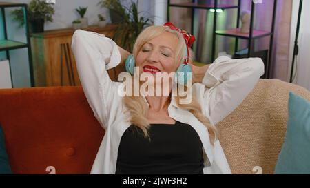 Felice e rilassata donna anziana gioita in cuffie wireless sul divano a casa scegliendo l'ascolto di musica rock and roll discoteca preferita energetica da app per smartphone divertente. Persone attività weekend Foto Stock