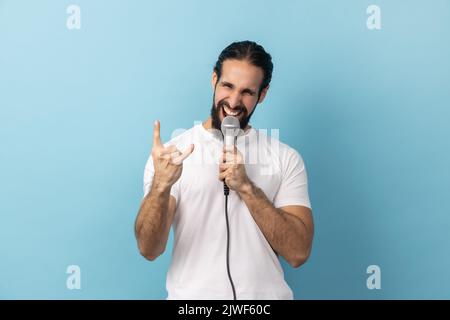 Ritratto di un uomo eccitato con barba in T-shirt bianca cantando forte canzone tenendo in mano il microfono, divertendosi a riposare nel karaoke, mostrando segno rock. Studio in interni isolato su sfondo blu. Foto Stock