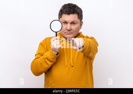 Detective serio e sicuro dell'uomo che tiene in mano la lente d'ingrandimento e punta il dito su di te guardando la fotocamera, spioncendo, indossando felpa con cappuccio stile urbano. Studio in interni isolato su sfondo bianco. Foto Stock