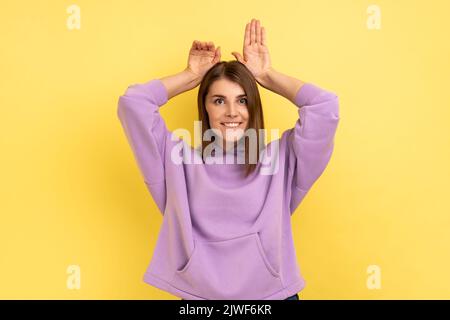 Ritratto di divertente spensierata attraente donna che mostra coniglietto orecchie gesto e aspetto allegro, comportamento infantile, con cappuccio viola. Studio al coperto isolato su sfondo giallo. Foto Stock